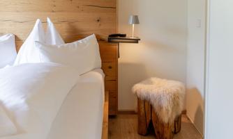 A cozy bedroom with a wooden bed and white pillows. Next to the bed is a wooden stool with a fur.