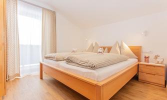 A cozy bedroom with a large bed and two pillows. The windows provide plenty of daylight and the wood details give the room a warm character.