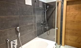 A modern bathroom with a bathtub and a shower. The walls are finished with dark tiles, and there is a wooden door.