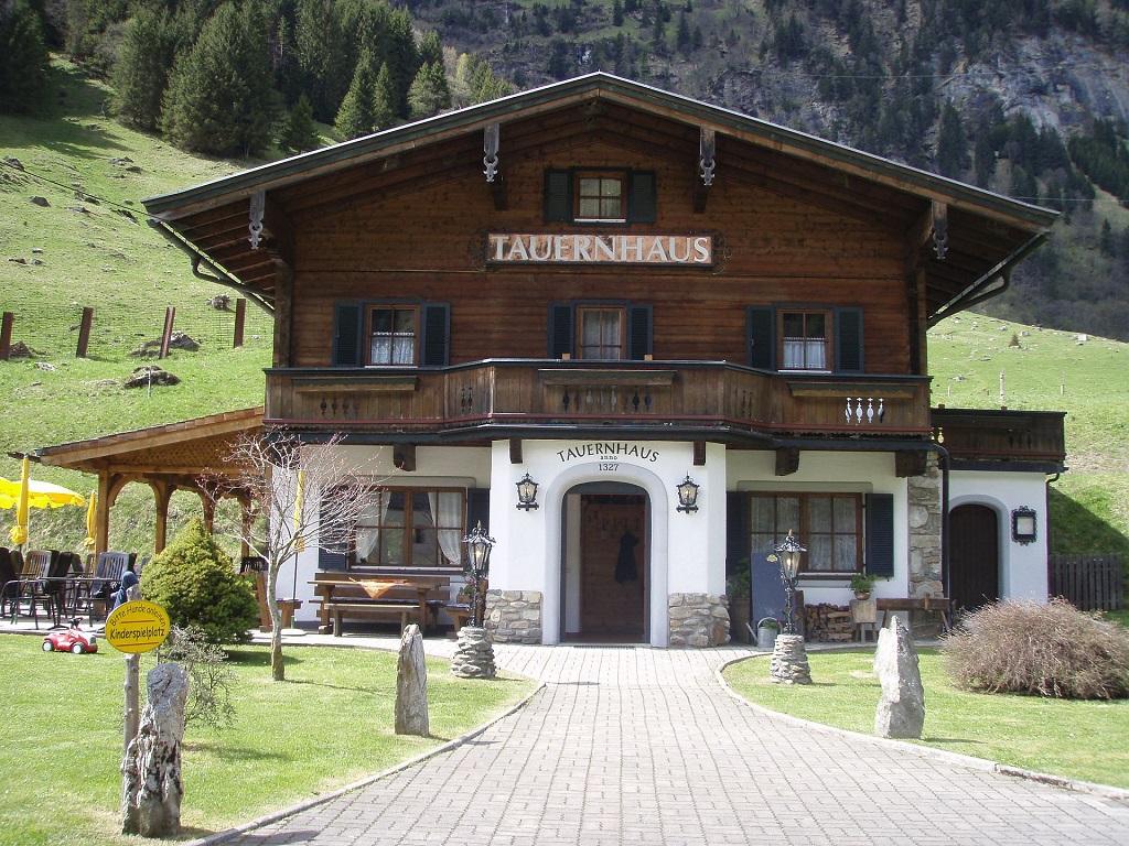 A traditional mountain house with wooden cladding and a green garden. In the background, high mountains are visible, framing the idyllic landscape.