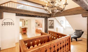 A bright and cozy interior with a wooden beam and an elegant chandelier. In the background, various rooms are visible, creating an inviting atmosphere.