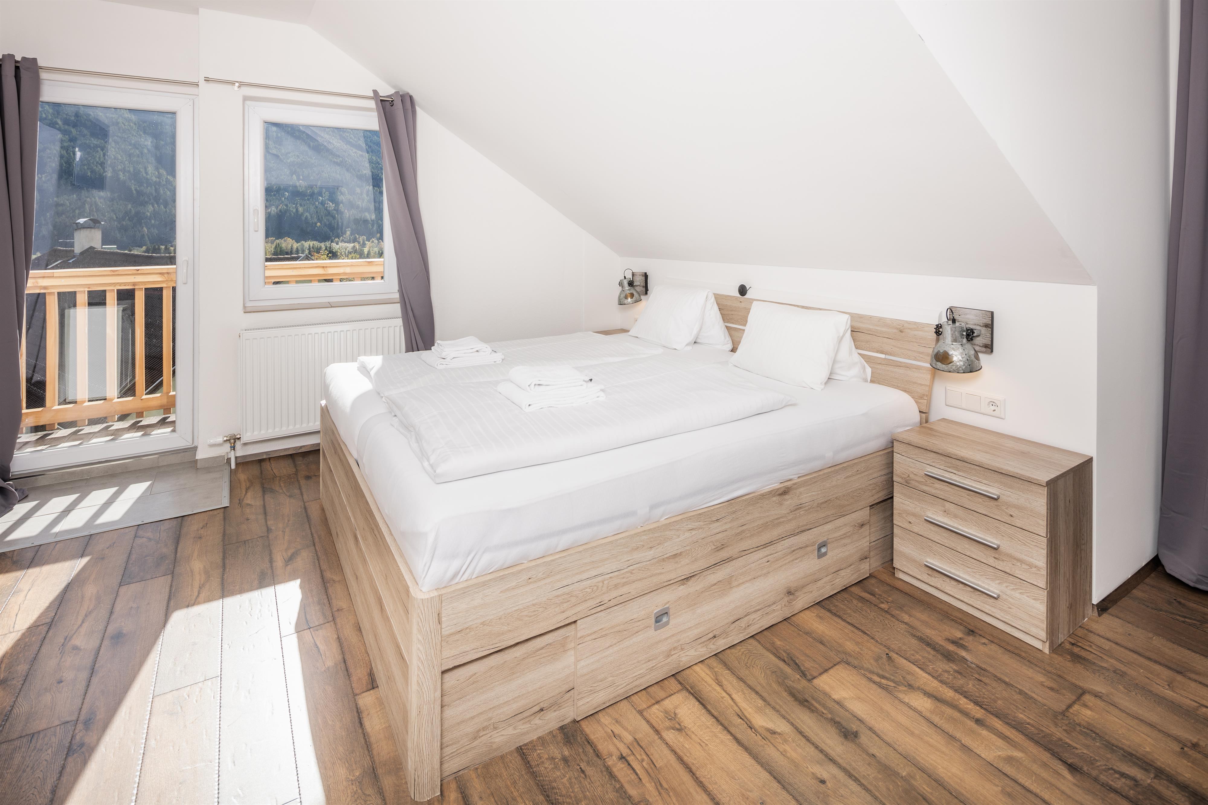 A bright bedroom with a large bed and wooden flooring. The windows offer a view of the mountains and let in plenty of daylight.