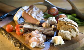 A rustic wooden board with various types of sausage and meat, fresh tomatoes, garlic, and bread. The arrangement conveys a hearty, traditional meal.