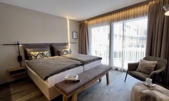 A modern bedroom with a large bed and two bedside tables.  
Next to the bed is a cozy armchair and a table, and large windows let in a lot of light.