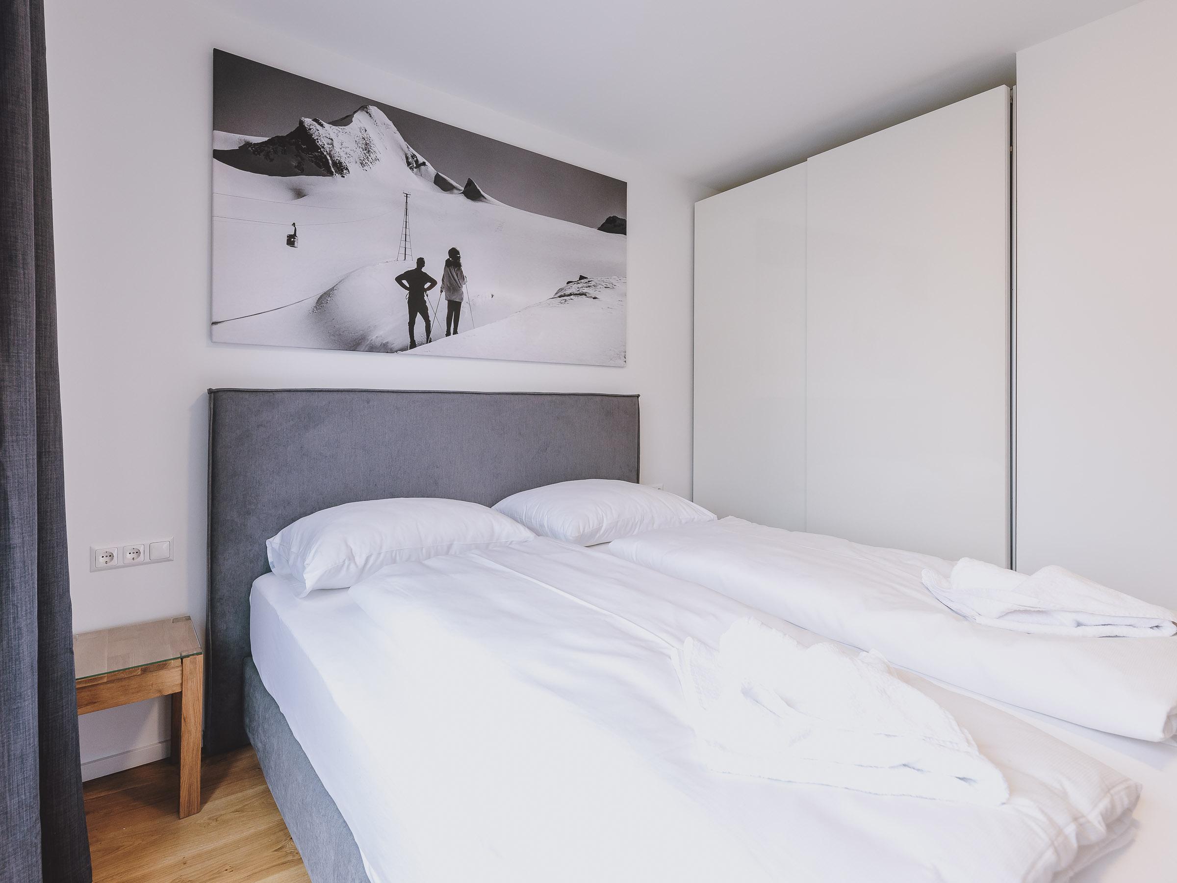 A modern bedroom with a double bed and white bed linens. On the wall hangs a picture of snow-covered mountains and people in the snow.