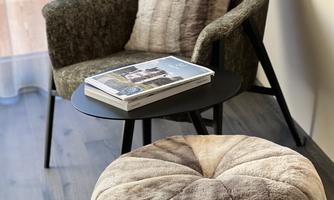 A cozy armchair with a matching cushion sits next to a small table. On the table lies a book and an elegant ottoman is also present.