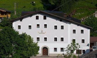 A white building with many windows and an entrance in the middle. Surrounded by trees and gentle hills in the background.