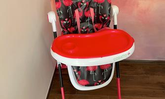 A high chair for children with a colorful flamingo pattern and a red table attachment. The chair is placed against a wall with a gentle gradient.