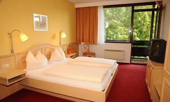 A bright and cozy bedroom with two single beds and modern wooden furniture. Large windows offer a view of the greenery and bring plenty of daylight into the room.