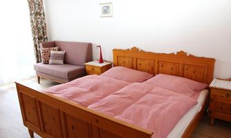 A cozy bedroom with a wooden bed and pink bedspreads. Next to the bed is a lilac couch and a small nightstand.
