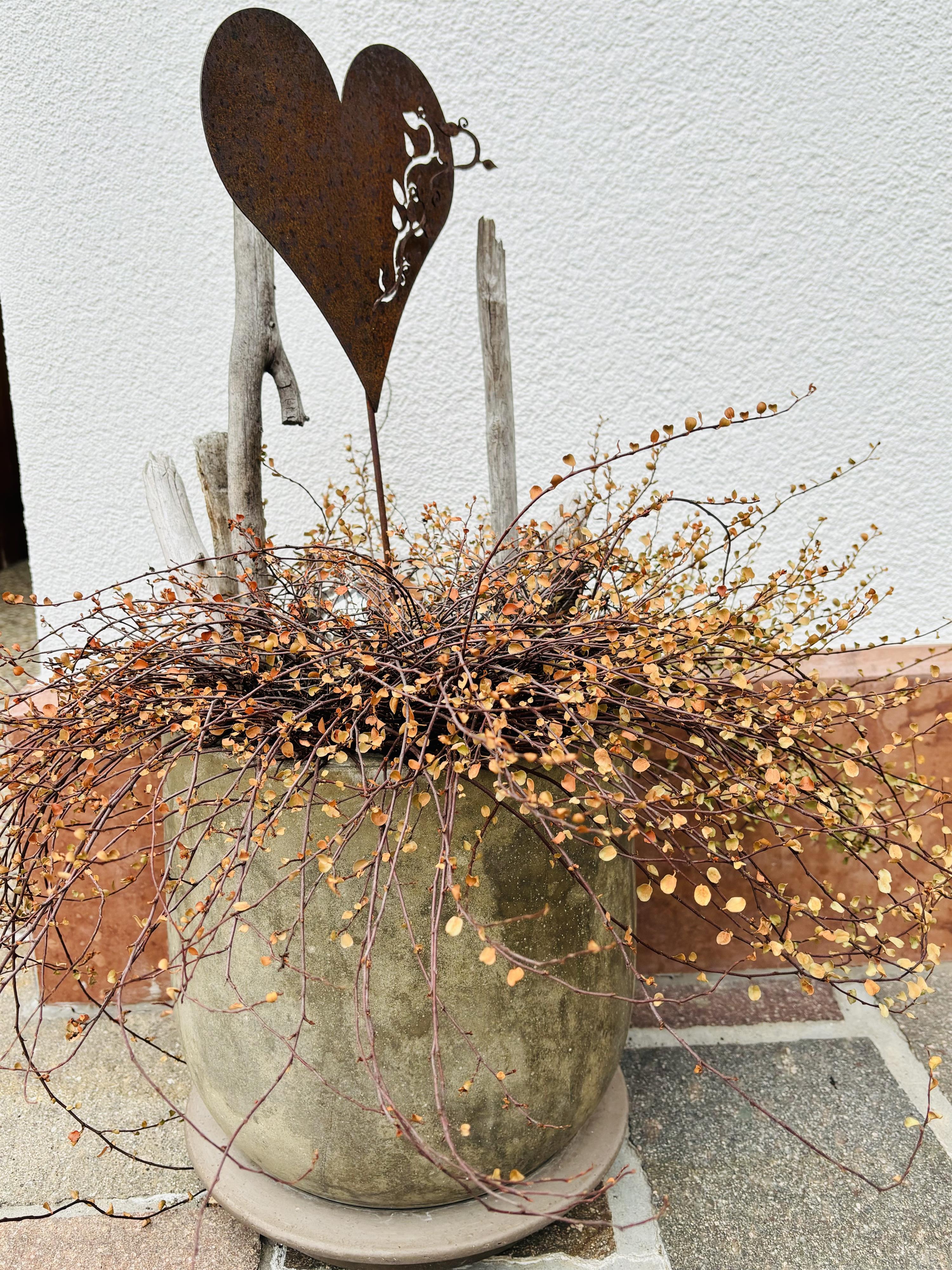 A blooming flower pot with dried branches and golden leaves. A heart-shaped metal sign complements the decorative arrangement.