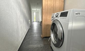 A narrow laundry room with a white washing machine and a wooden cabinet. The floor is tiled with dark plates, and there is plenty of light from an open space at the end.
