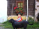 A small child with a hat sits next to a black dog in front of a house. Red blooming flowers are flourishing in the window.