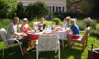 A group of five people is sitting at a table in the garden and enjoying breakfast. In the background, trees and a house are visible.