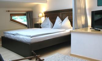 A cozy bedroom with a large bed and white pillows. The room is bright and furnished in a modern style, with a television and a pleasant view outside.