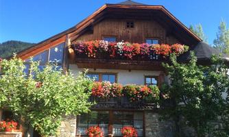 A beautiful house in alpine style with blooming balcony plants. In the foreground, green trees stand and a clear blue sky is visible.