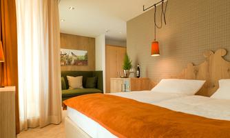A modern hotel room with a double bed and orange bedding. The room features large windows, bright curtains, and a cozy seating area.