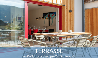 A bright terrace with a large table and chairs. In the background, there is a dining area with modern lamps.