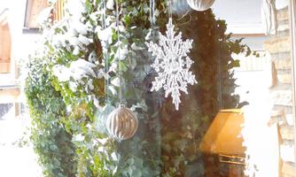 A cozy winter view with snow-covered plants and festive decorations. A sparkling snowflake and Christmas ornaments hang in front of a window.