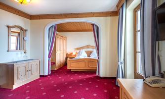 A cozy hotel room with wooden furniture and a red carpet. In the background, a bed with white bedding and a wardrobe are visible.