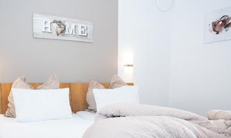 A bright bedroom with a comfortable double bed and decorative pillows. A sign with the word "HOME" hangs on the wall.