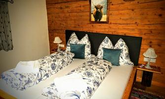 A cozy bedroom with a double bed decorated with decorative cushions and bedding. On the wall hangs a picture of a cow, and on the nightstands are small lamps.
