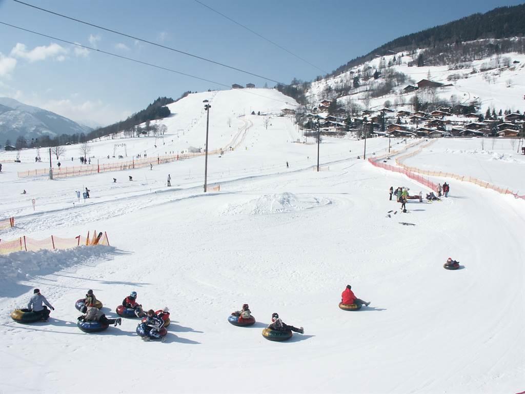 A snow-covered slope with cheerful people tubing. In the background, other skiers and a clear blue sky can be seen.