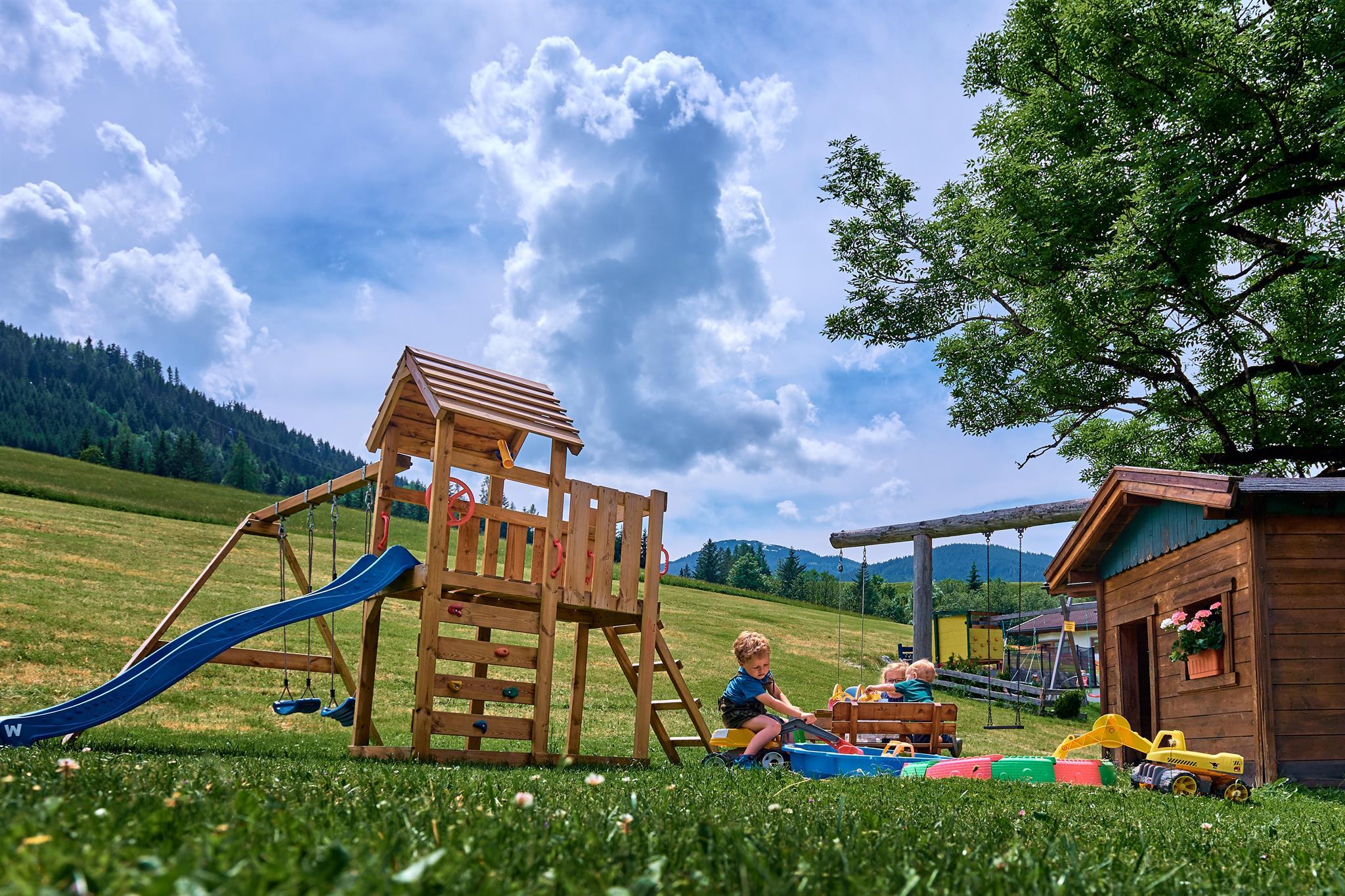 A playground with a wooden slide and a play tower construction. In the background, a green field and some clouds can be seen.