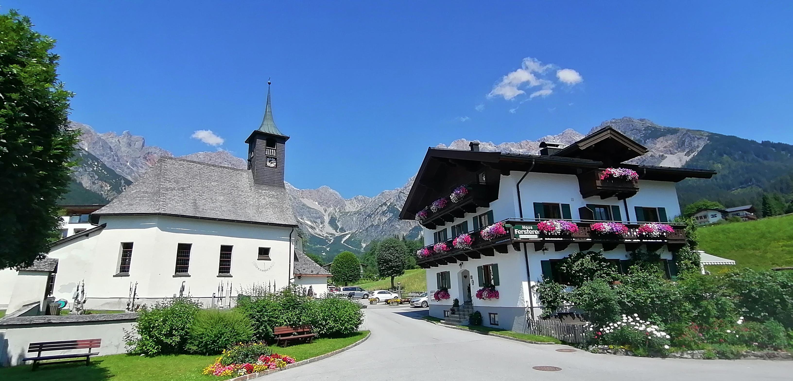 A picturesque village scene with a church and a traditional house. The surroundings are surrounded by green meadows and mountains, under a clear blue sky.