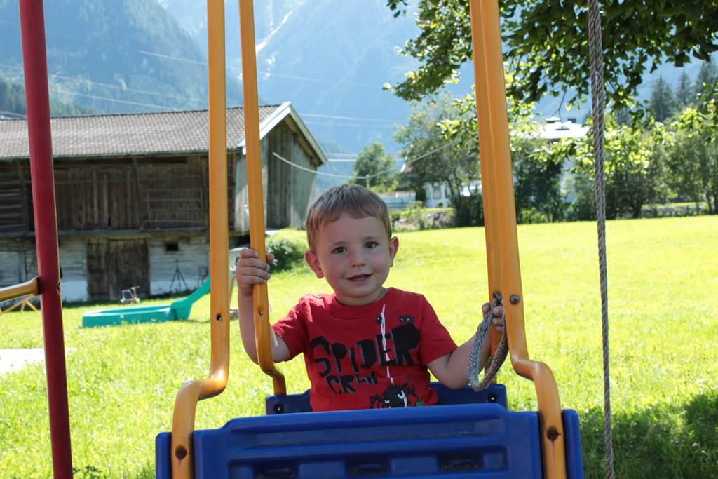 A little boy is sitting on a swing and smiling. In the background, green meadows and mountains can be seen.