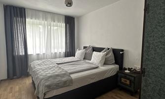 A modern bedroom with a double bed and windows with curtains. The wooden floor and minimalist furnishings create a cozy atmosphere.