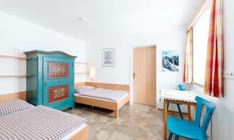 A simple room with two single beds and a colorful wardrobe. There is a small table with a blue chair and plenty of natural light.
