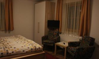 A simple bedroom with a bed, two upholstered chairs, and a small table. The walls are light, and the curtains are golden.
