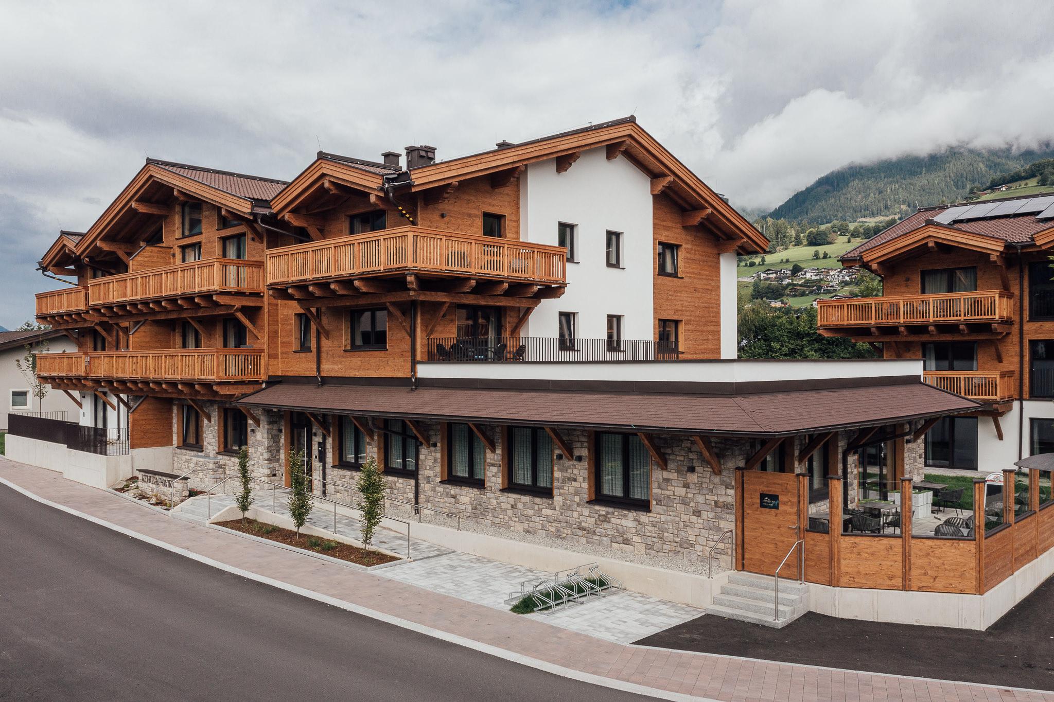 A modern building made of wood and stone with several balconies. In the background are gentle hills and a cloudy sky.
