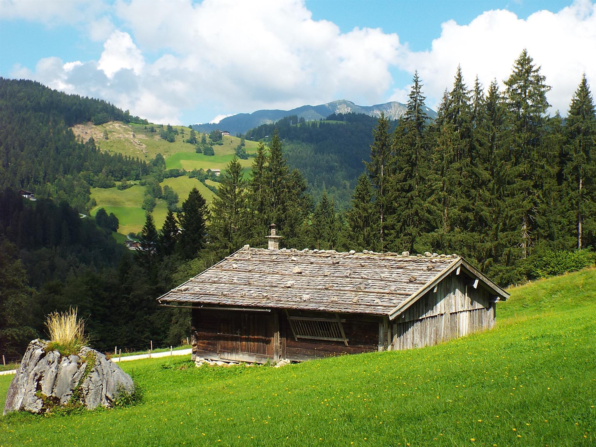 A cozy cottage in a green meadow, surrounded by trees and mountains. The clouds in the blue sky give the landscape a relaxed atmosphere.