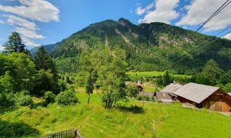A beautiful mountain landscape with green meadows and a clear sky. In the foreground, there are trees and some huts.