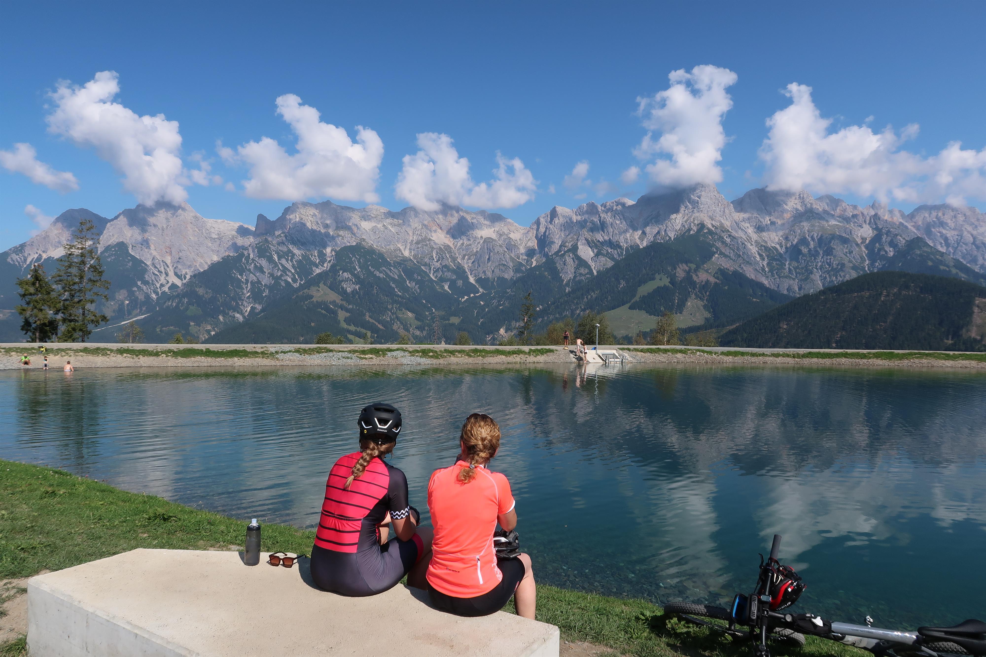 Two people are sitting by the shore of a clear lake, enjoying the view of the mountains. The sky is blue with some clouds.