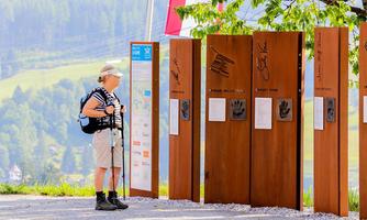 A hiker is standing at an information point outdoors. In the background, the mountains and a clear sky are visible.