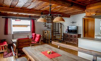 A cozy living space with a wooden ceiling and rustic furniture. The room has a staircase and large windows with red curtains.
