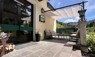 An inviting outdoor area with comfortable seating and shade-providing sails. In the background, green hills and a blue sky can be seen.