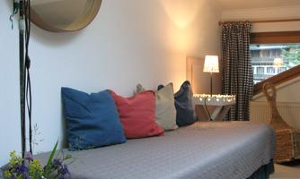 A cozy room with a sofa bed, colorful cushions, and soft lighting. A large mirror and a table with a small flower vase complete the atmosphere.