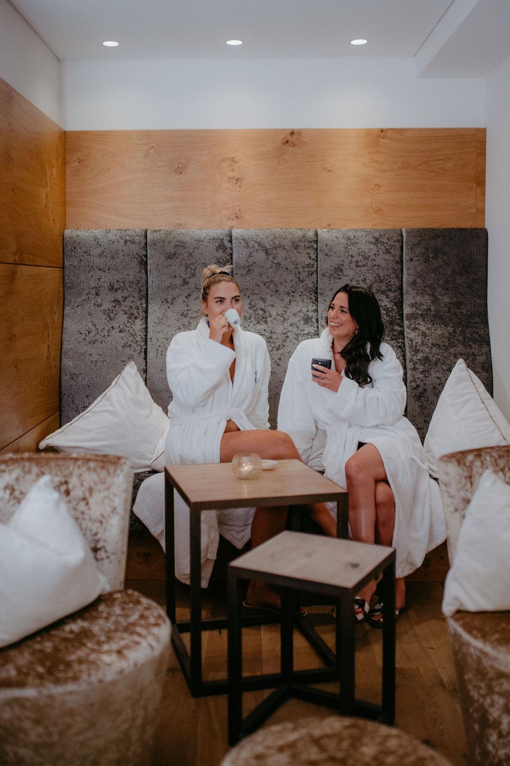 Two women in bathrobes relax in an elegant wellness area. They enjoy drinks in a cozy atmosphere.