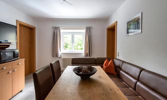 A bright and modern dining area with a wooden table and a leather bench. In the background are windows and a small kitchen.
