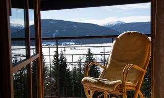 A comfortable rocking chair stands in front of a large window with a view of snow-covered mountains and forests. The landscape radiates tranquility and idyllic charm.