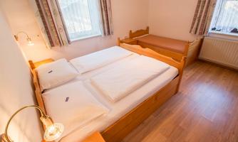 A cozy bedroom with two beds and warm wooden decor. The room has windows with curtains and a bright wooden floor.