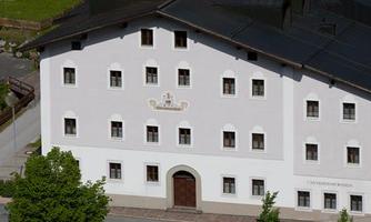 A large, white building with several windows and a central entrance. Around the building, there are trees and additional small structures.