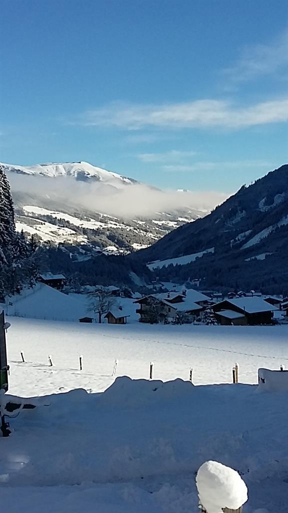 A winter landscape with snow-covered mountains and a clear blue sky. In the foreground, there are snowy meadows and scattered houses.