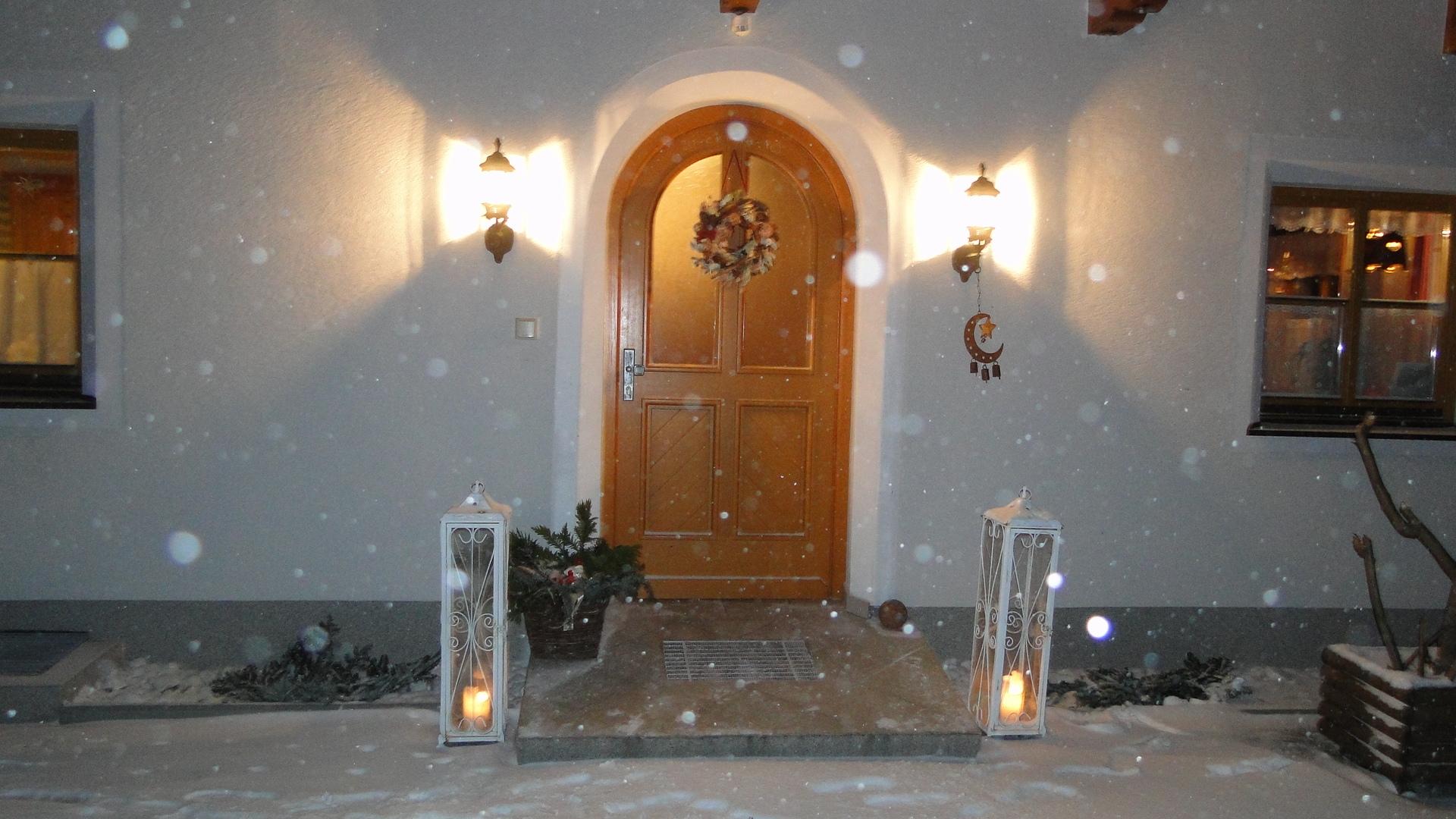 A cozy front door with a Christmas wreath, surrounded by snow-covered ground. The lights at the entrance create a warm atmosphere in the cold winter night.