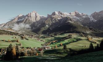 A picturesque landscape with high mountains in the background. In the foreground, there are green meadows and small houses.