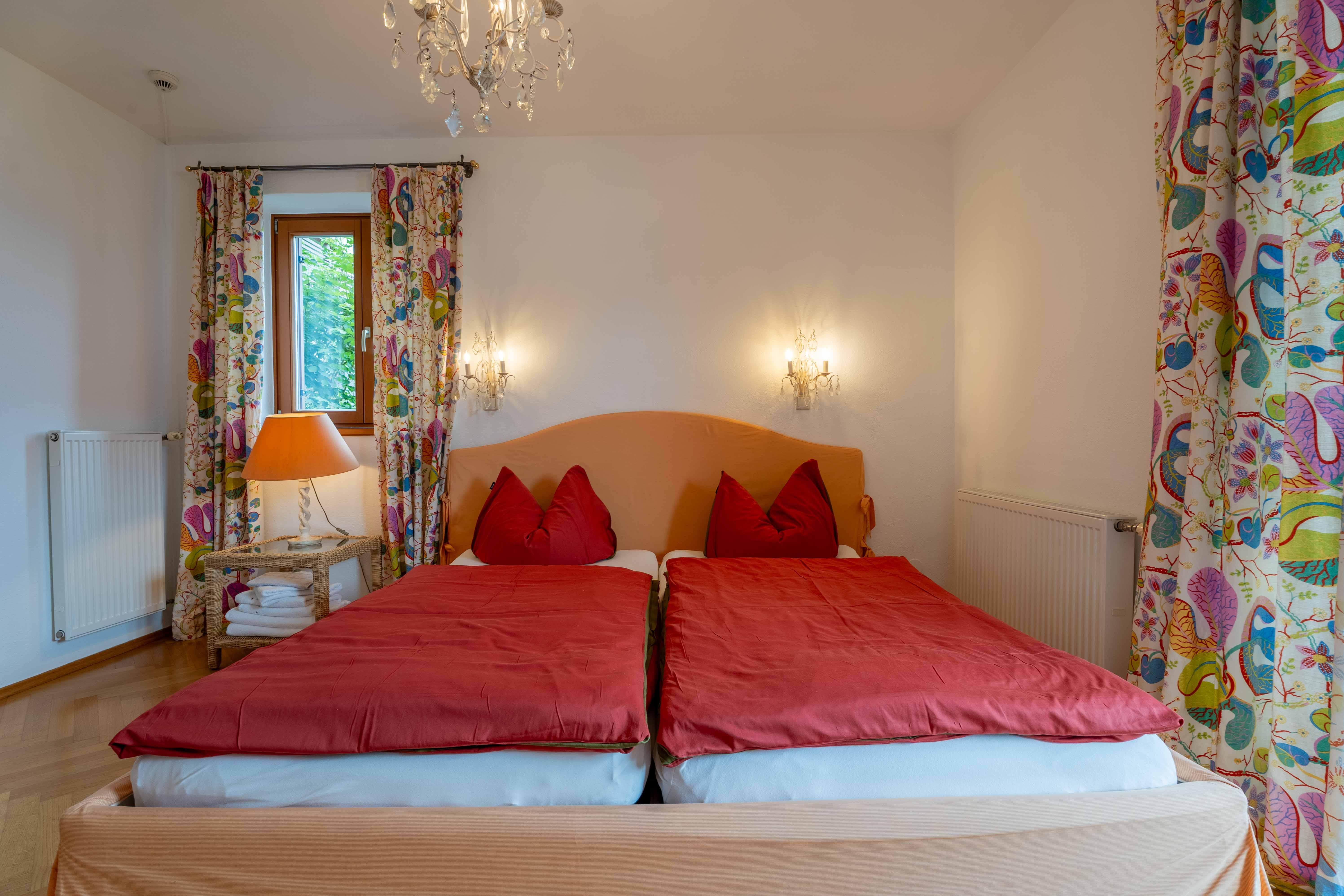 A cozy bedroom with a large bed and red bedspreads. Colorful curtains and a lamp give the room an inviting atmosphere.